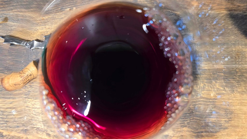 Red wine sitting in the bottle of a clear glass on a wood countertop.